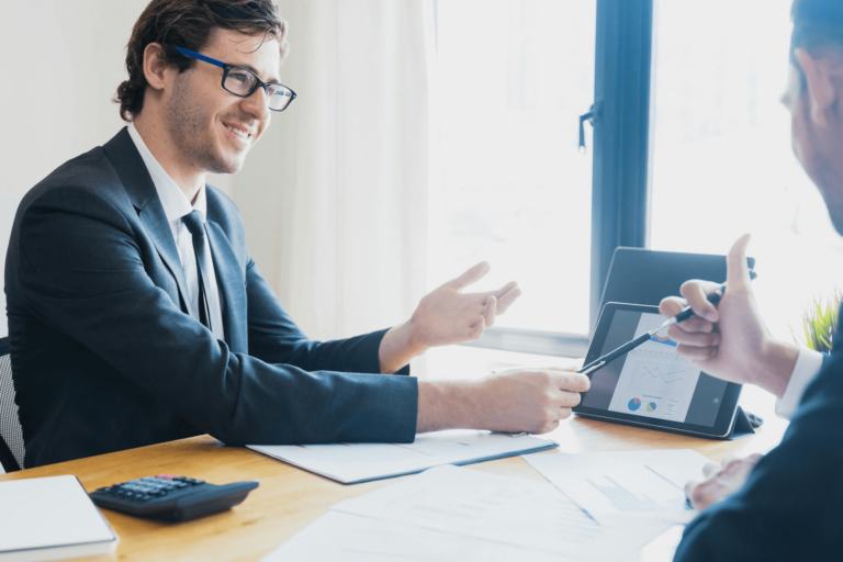 Two businesspeople in suits are having a meeting at a desk with documents, and a tablet.